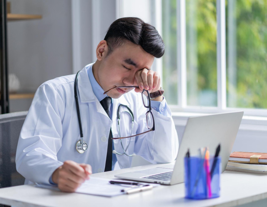 Healthcare provider sitting at a desk looking fatigued while reviewing work on a laptop, representing burnout and career stress