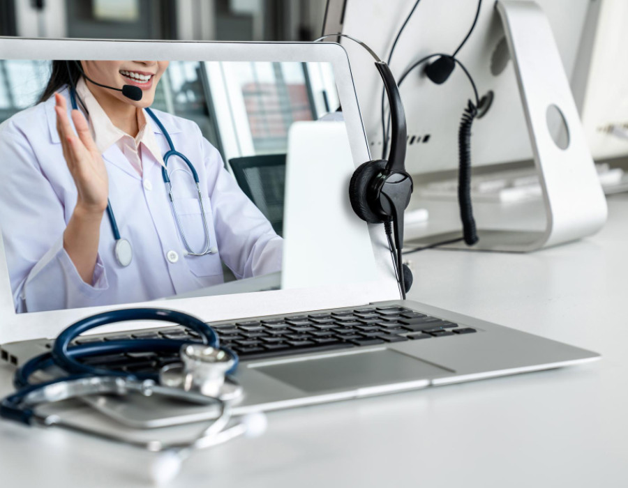 Doctor conducting a telemedicine visit on a laptop, wearing a headset and stethoscope, with medical equipment on a desk.