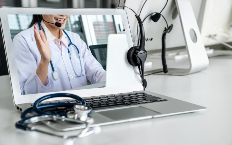 Doctor conducting a telemedicine visit on a laptop, wearing a headset and stethoscope, with medical equipment on a desk.