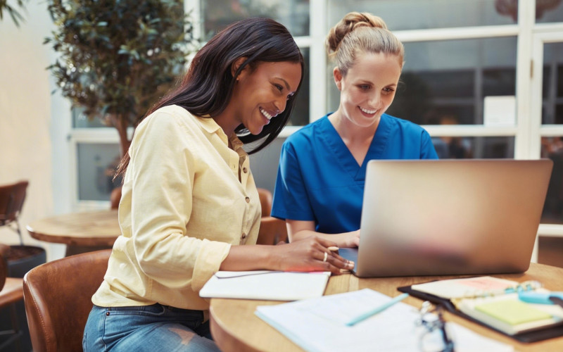 Nurse in scrubs collaborating on a laptop with a colleague, representing tutoring and remote side hustle opportunities for nurses