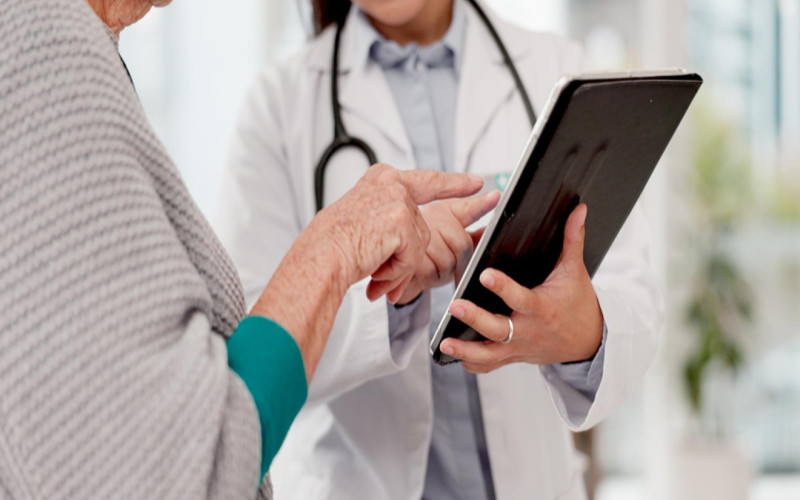 Nurse practitioner reviewing patient information on a tablet while consulting with an elderly patient in a clinic setting.